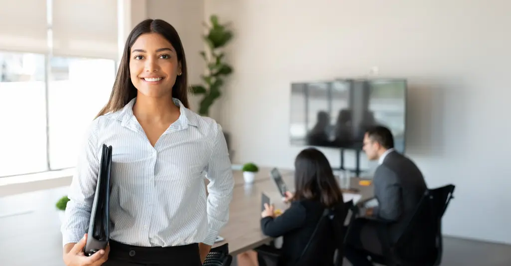 Young professional standing in front of conference room while holding a binder.